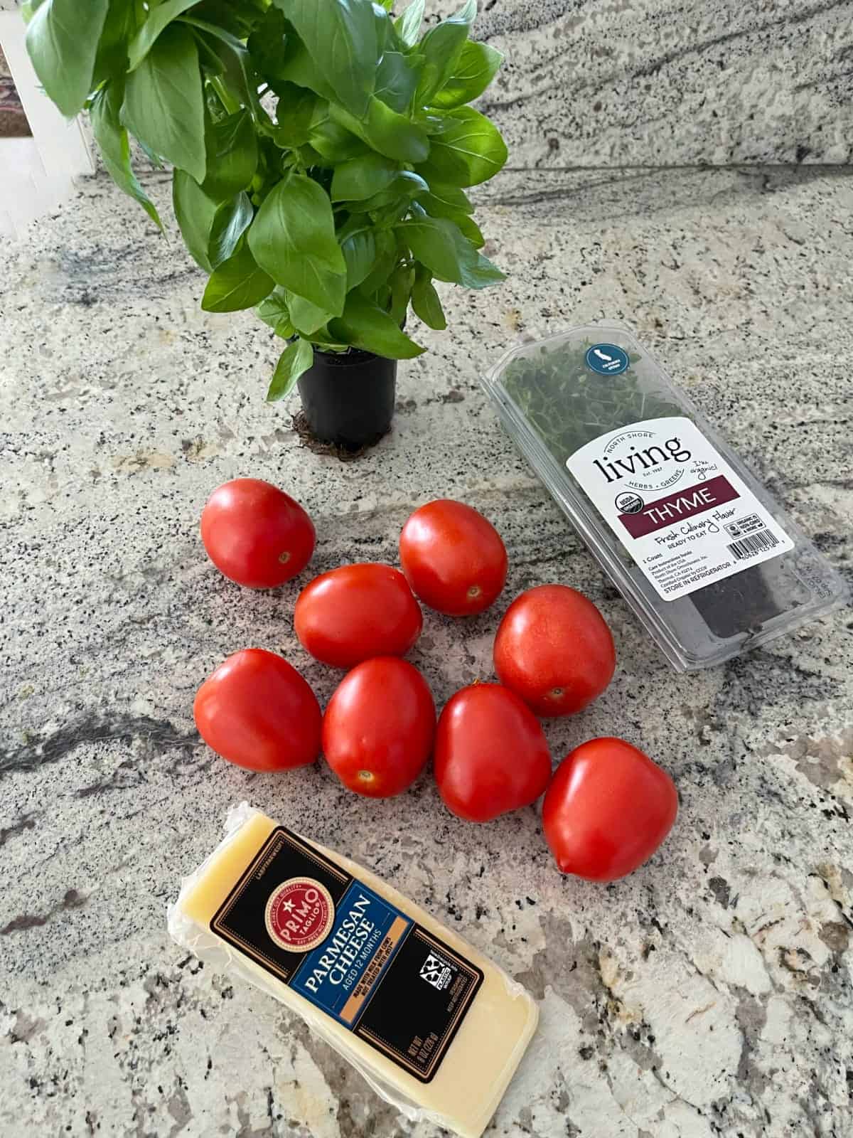 Ingredients including basil plant, package of fresh thyme, Romat tomatoes and Parmesan cheese on granite.