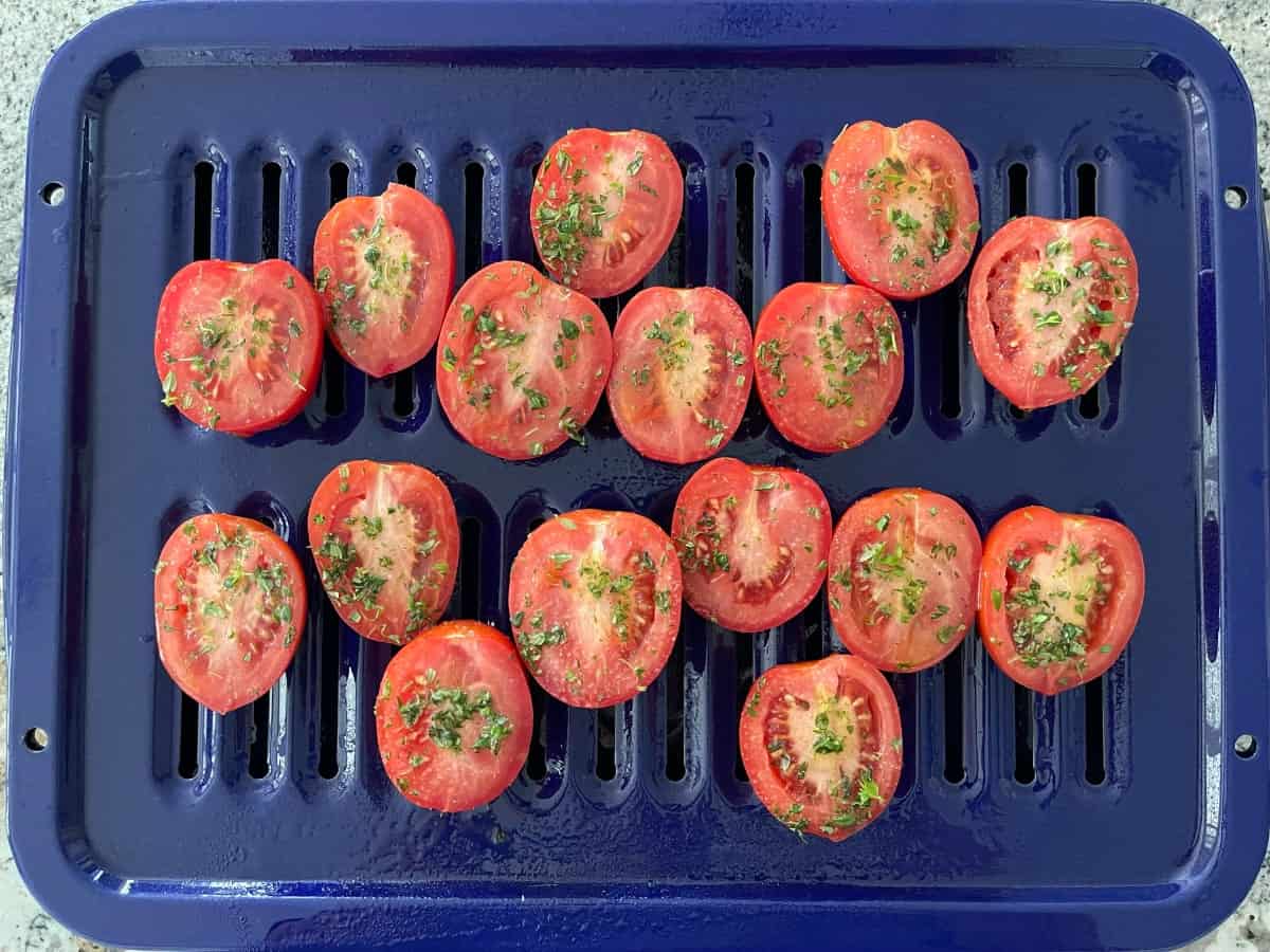 Halved Roma tomatoes tossed in olive oil, thyme, salt and pepper on blue broiler pan.