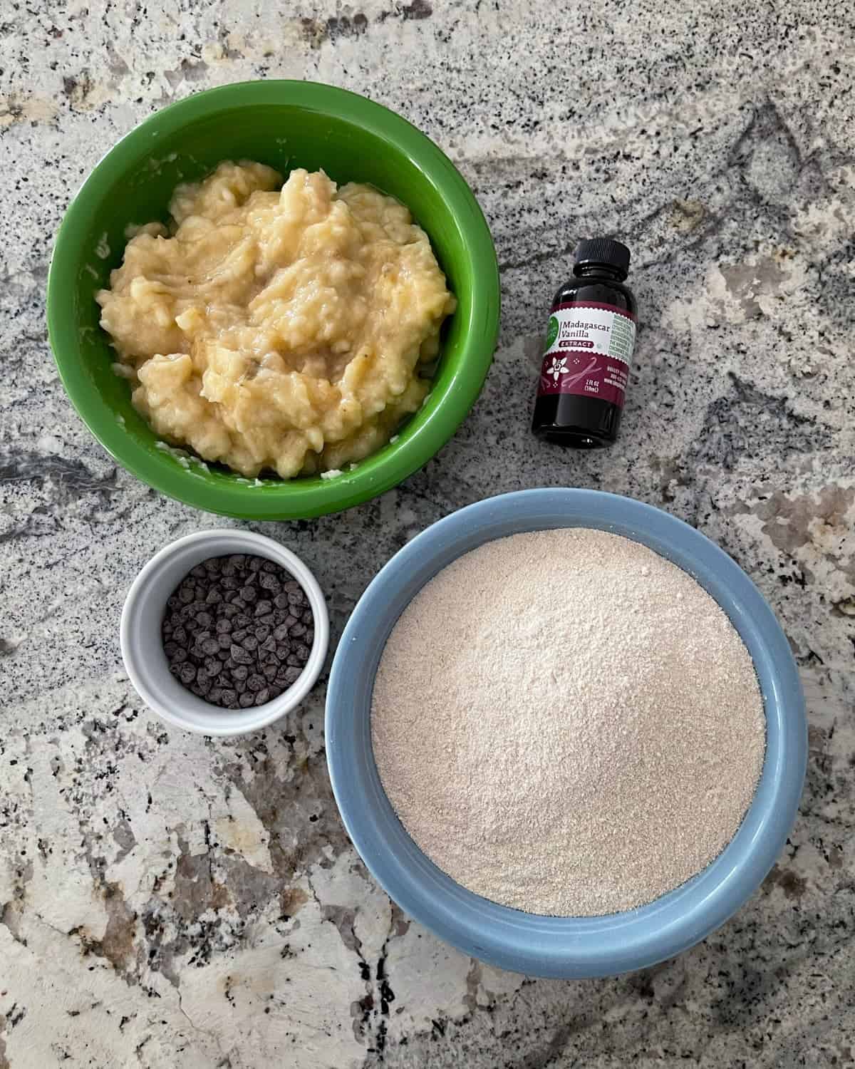Ingredients including mashed banana in green bowl, Kodiak Cakes flapjack mix in blue bowl, vanilla extract and miniature chocolate chips on granite.