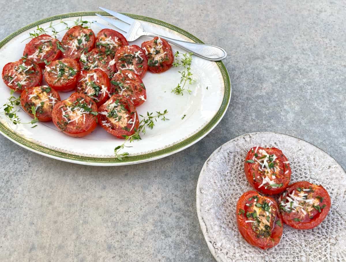 Parmesan broiled tomatoes on serving platter with large fork behind small plate with three broiled tomato halves.