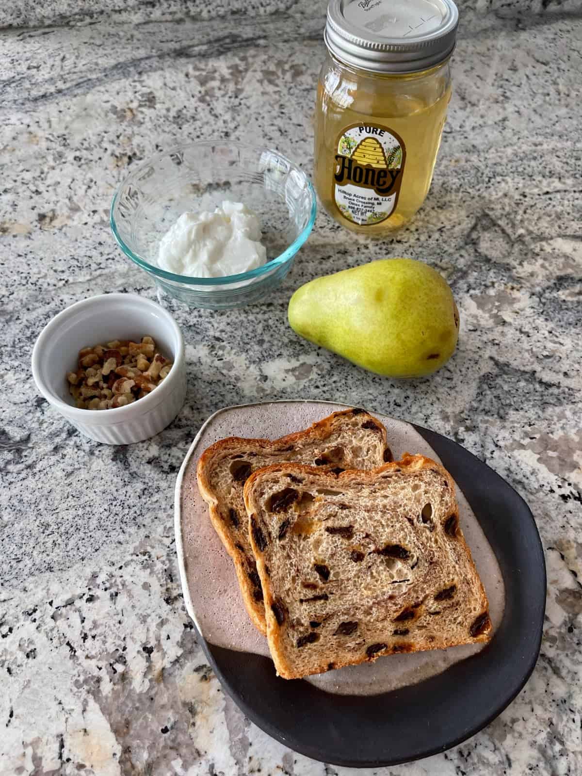 Ingredients including cinnamon raisin swirl bread, chopped walnuts, nonfat Greek yogurt, honey and a whole pear on granite counter.