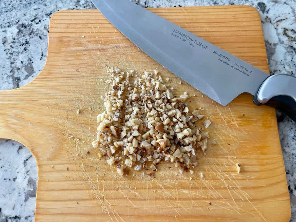 Finely chopping toasted walnuts on wood cutting board with chef's knife.