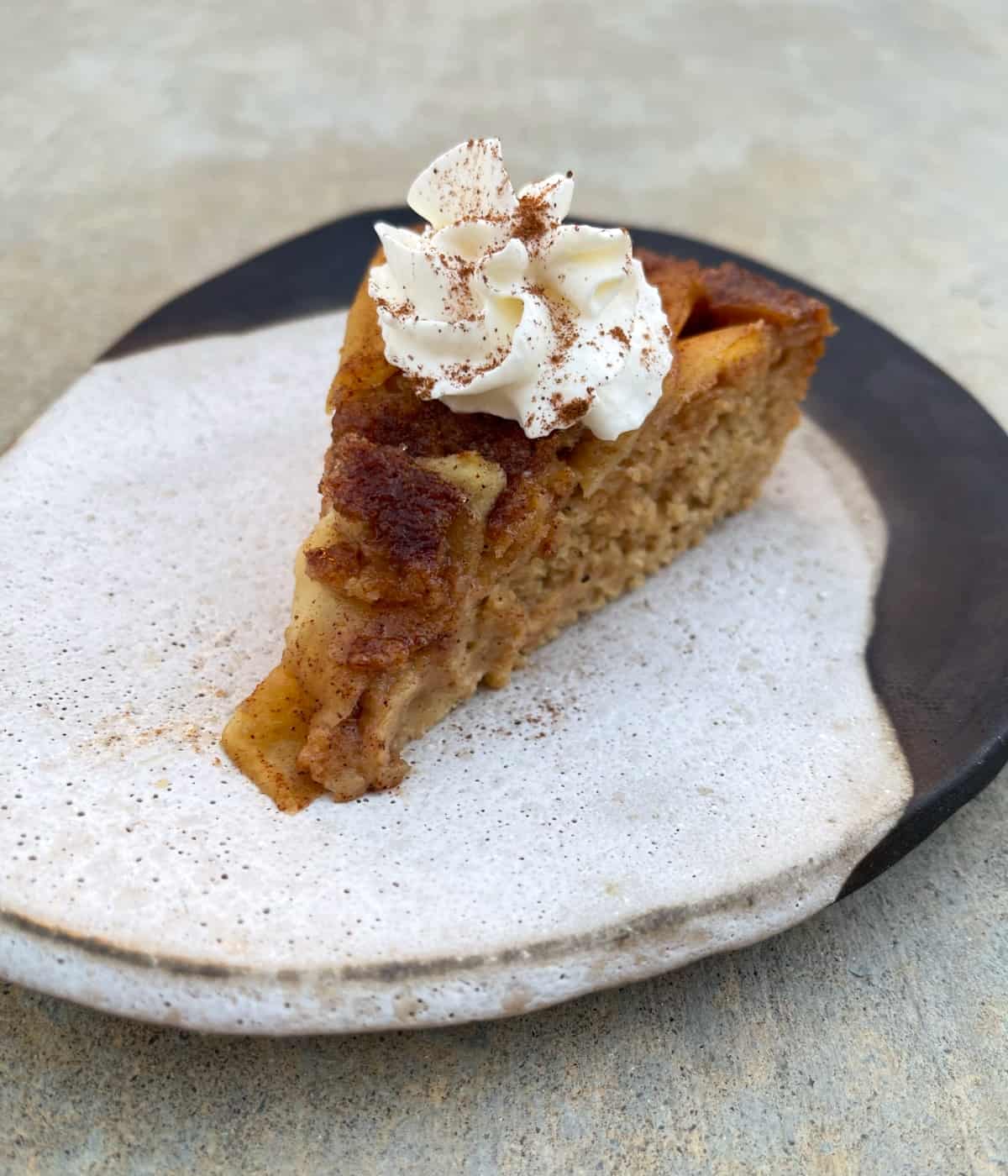 Slice of cinnamon apple quick cake garnished with whipped topping and sprinkle of cinnamon on ceramic plate.