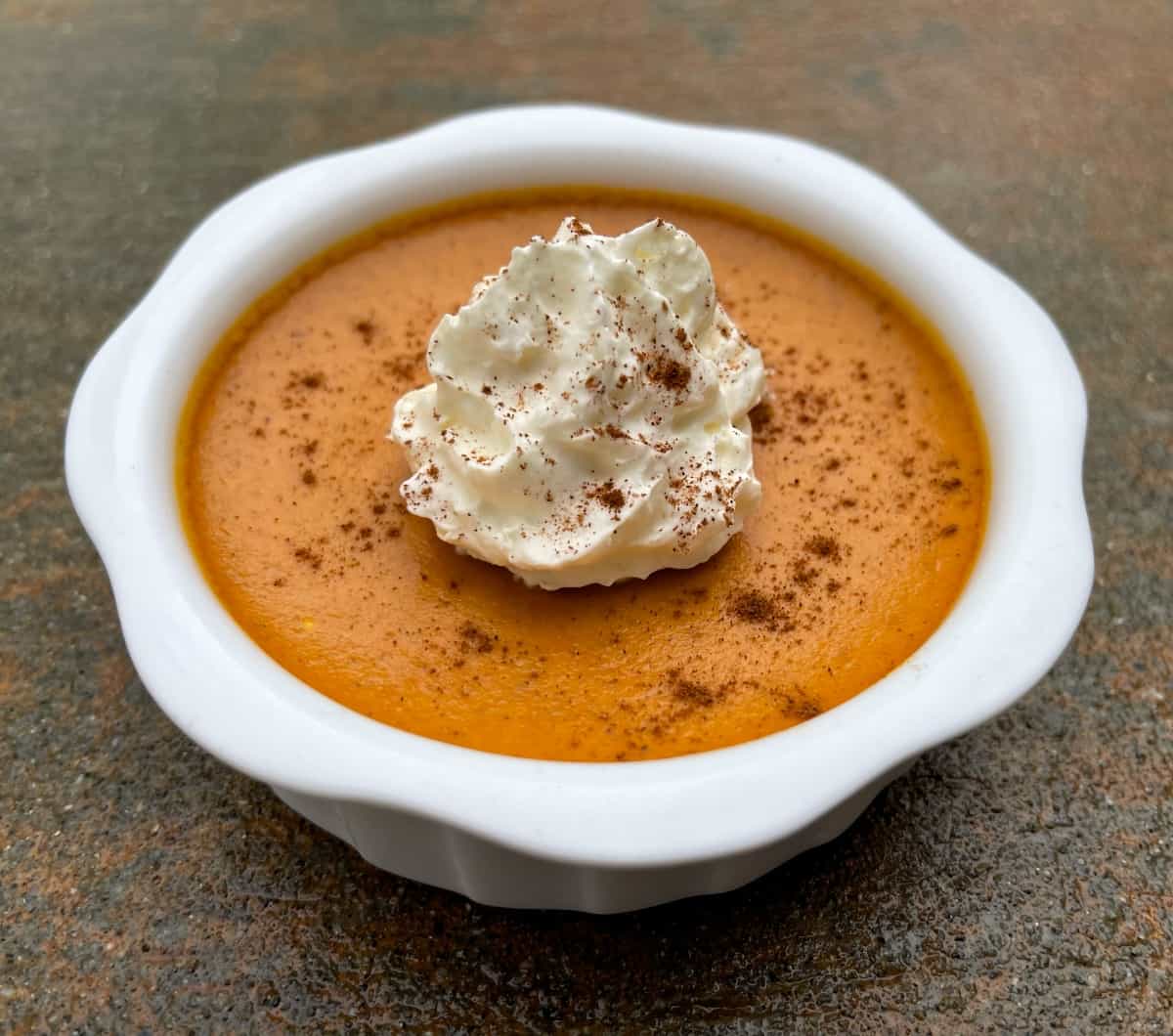 Individual creamy pumpkin pie custard cup topped with light whipped cream and sprinkle of cinnamon in white ramekin.
