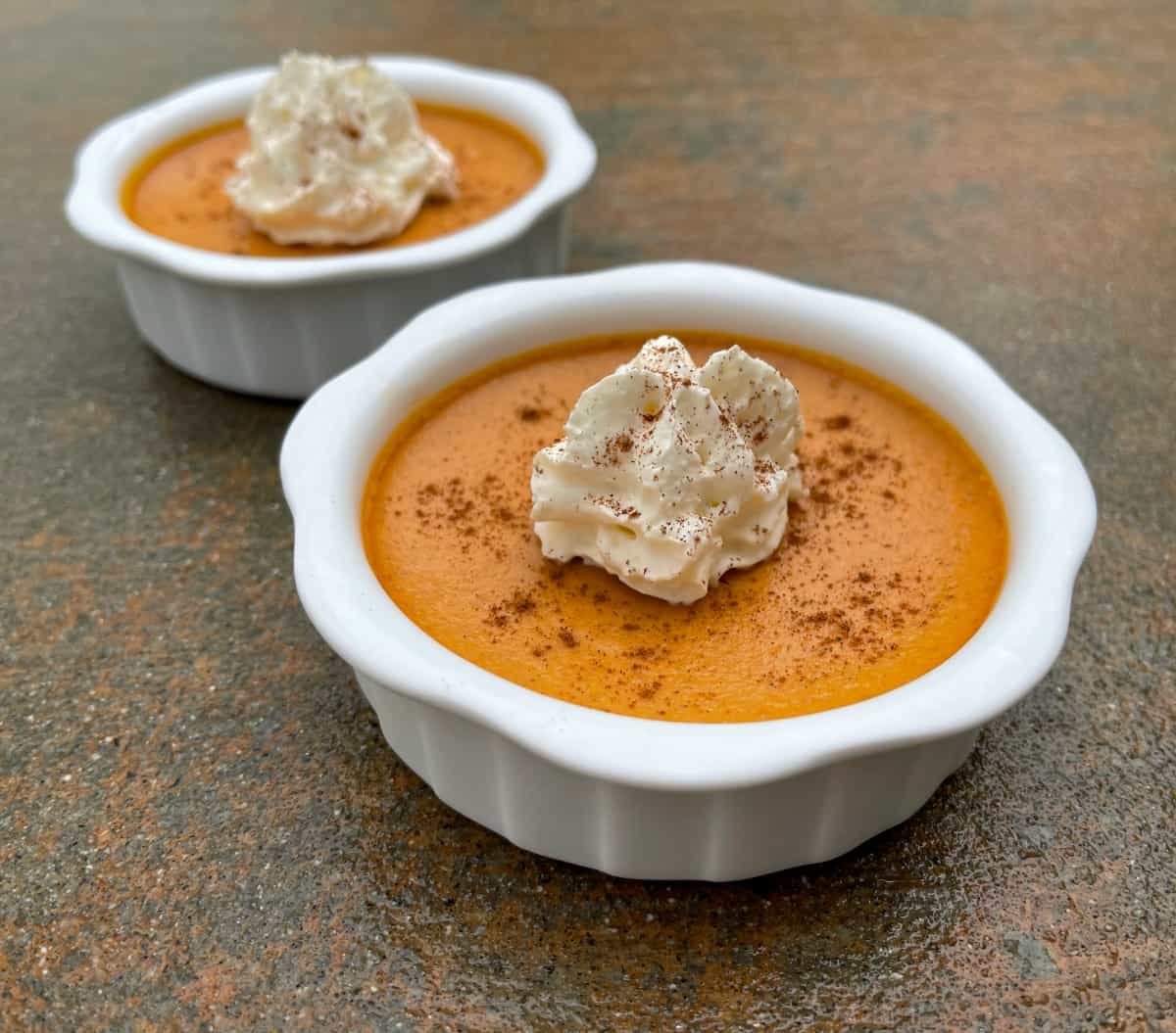 Super creamy pumpkin pie custard garnished with light whipped topping and sprinkle of pumpkin pie spice in two small white ramekins.