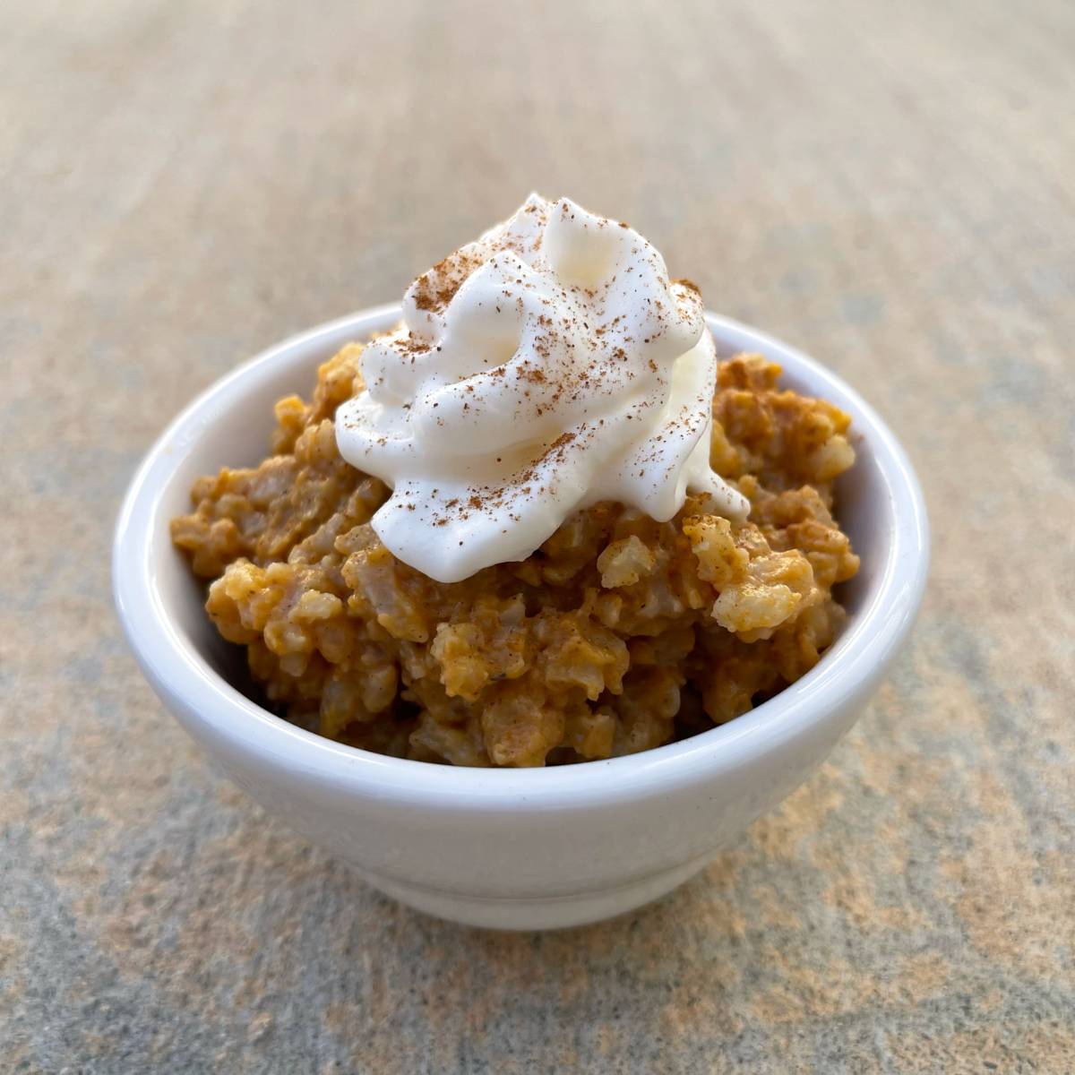 Pumpkin rice pudding in small white bowl topped with light whipped topping and cinnamon sprinkle.