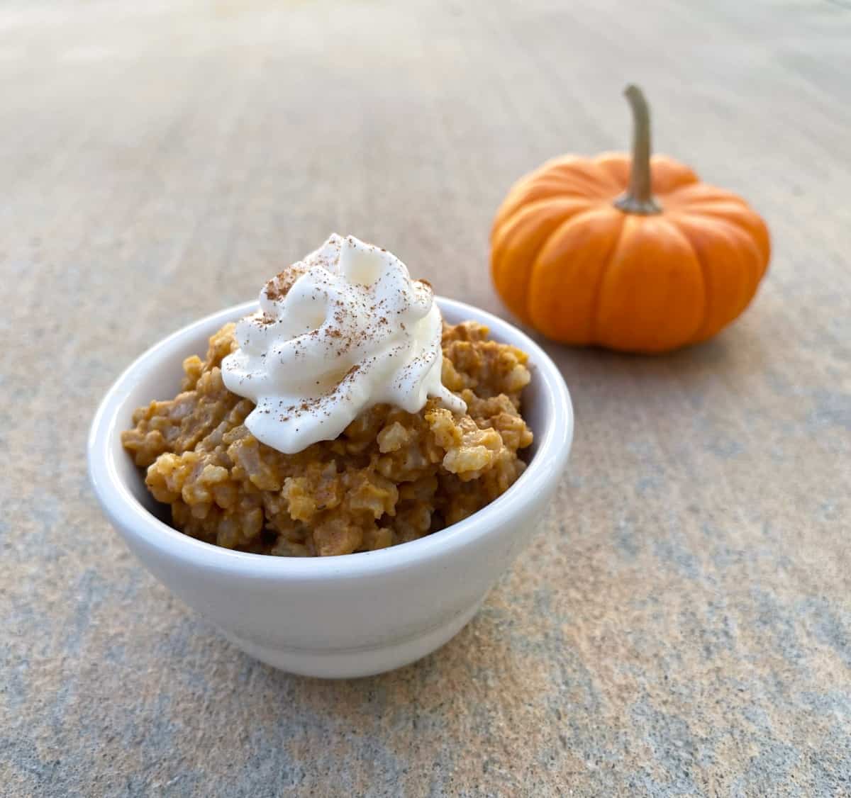 Pumpkin pie rice pudding in small white bowl topped with light whipped topping and cinnamon sprinkle with small orange pumpkin in background.