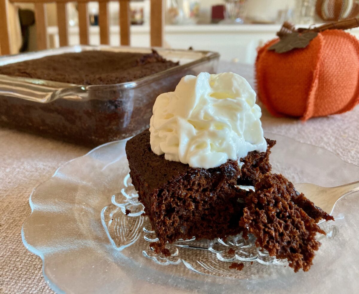 Piece of double ginger chocolate gingerbread topped with whipped cream with pan of gingerbread and decorative pumpkin in background.