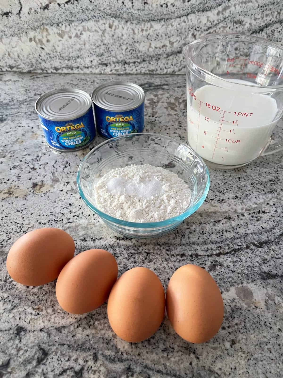 Ingredients including four eggs, two cans green chiles, unsweetened almond milk, and small glass bowl with all-purpose flour on granite.
