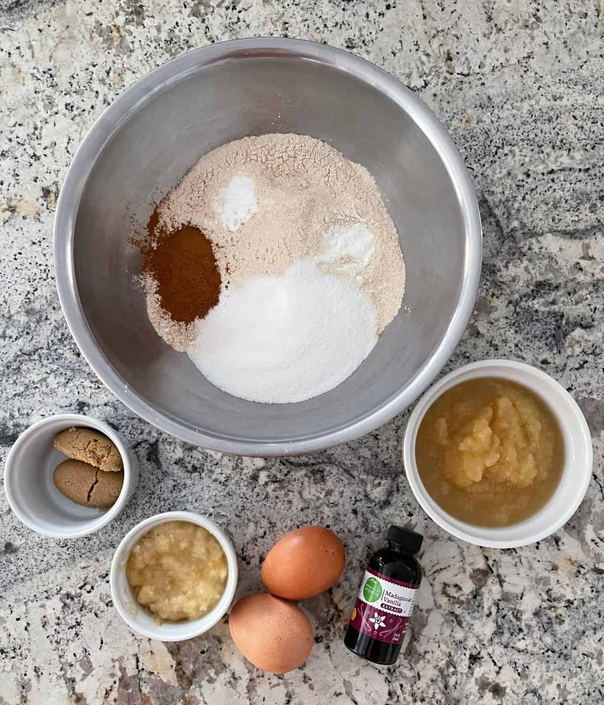 Muffin ingredients including a large mixing bowl with all-purpose flour, white whole wheat flour, cinnamon, Truvia Baking Blend, baking powder and baking soda, a small ramekin with mashed banana, unsweetened applesauce, 2 eggs, brown sugar and vanilla extract.