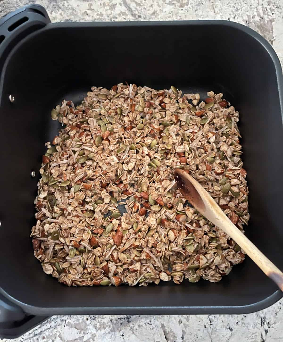Stirring oat granola in air fryer basket with wooden spoon halfway through cooking time.
