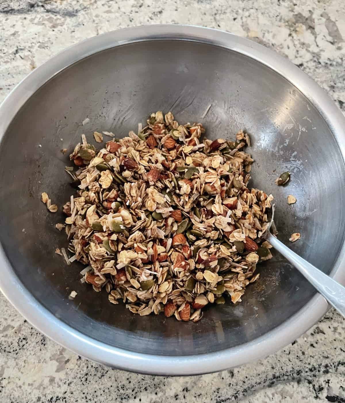 Stirring together rolled oats, chopped almonds, shredded coconut, pumpkin seeds (pepitas), coconut oil, maple syrup, ground cinnamon and cardamom in a mixing bowl with spoon.