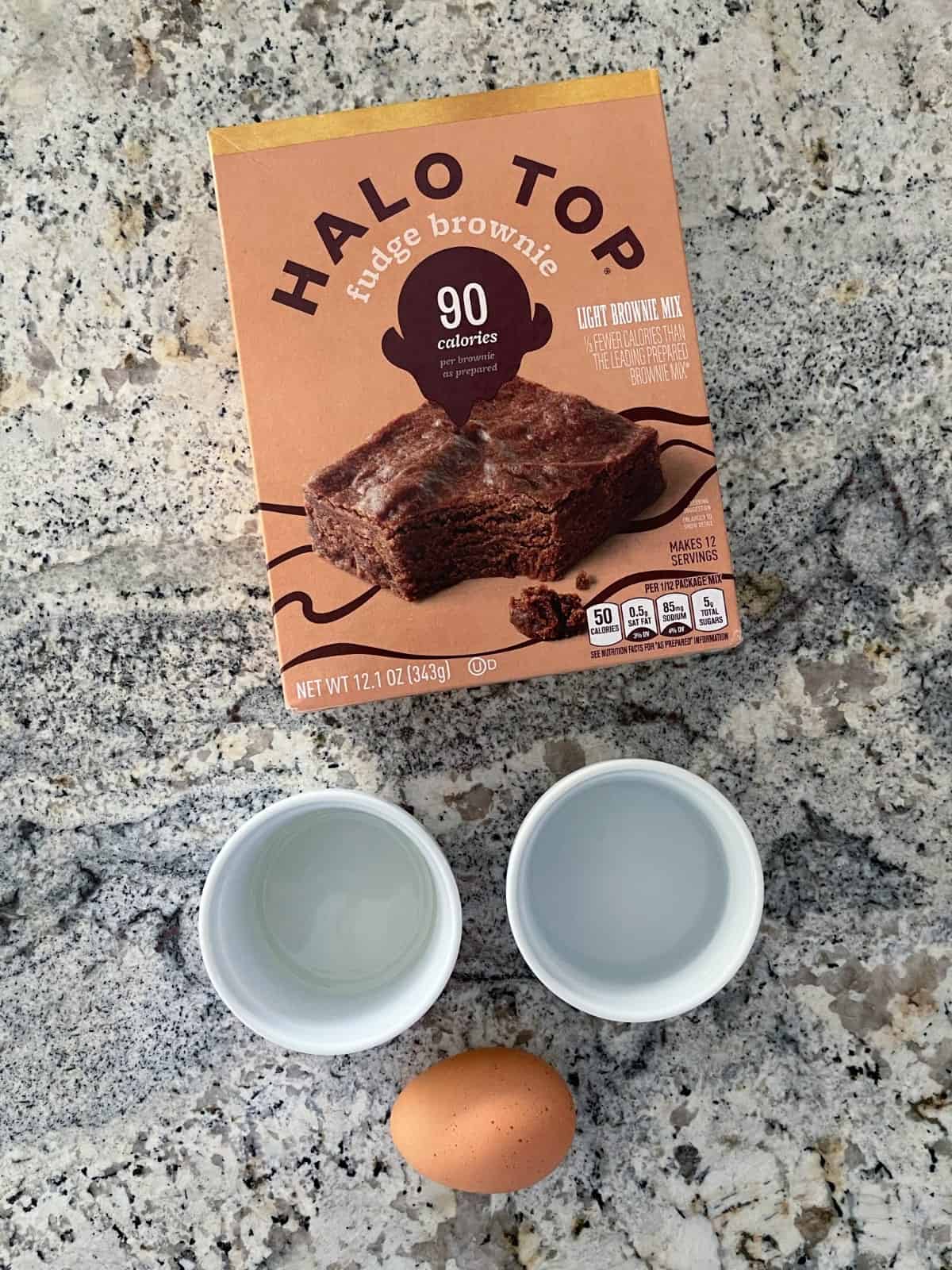 Box of Halo Top fudge brownie mix, two small white ramekins with water and canola oil and one egg on granite.