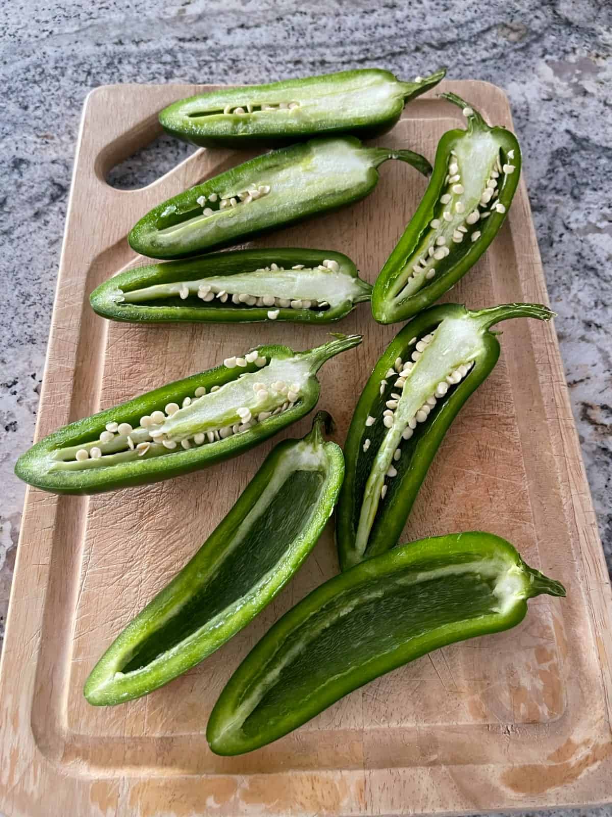 Jalapeno peppers on wood cutting board, cut in half lengthwise with ribs and seeds removed from two peppers.