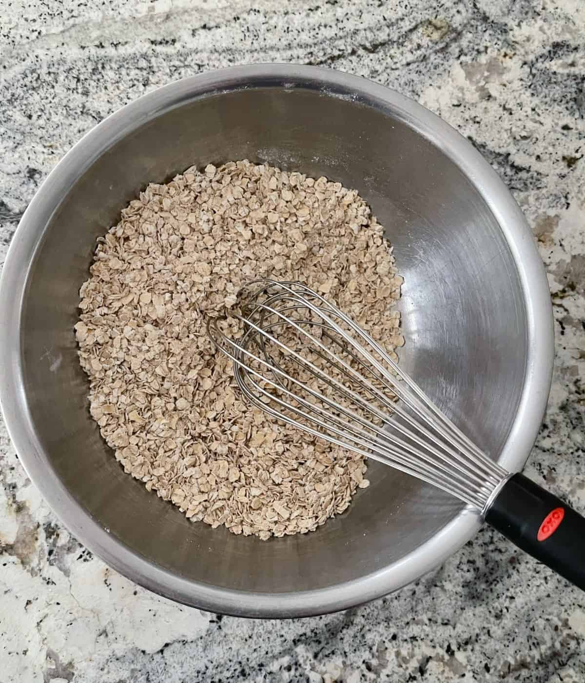 Mixing bowl with oats, flour, baking powder, baking soda, ground ginger and ground cinnamon and a whisk.