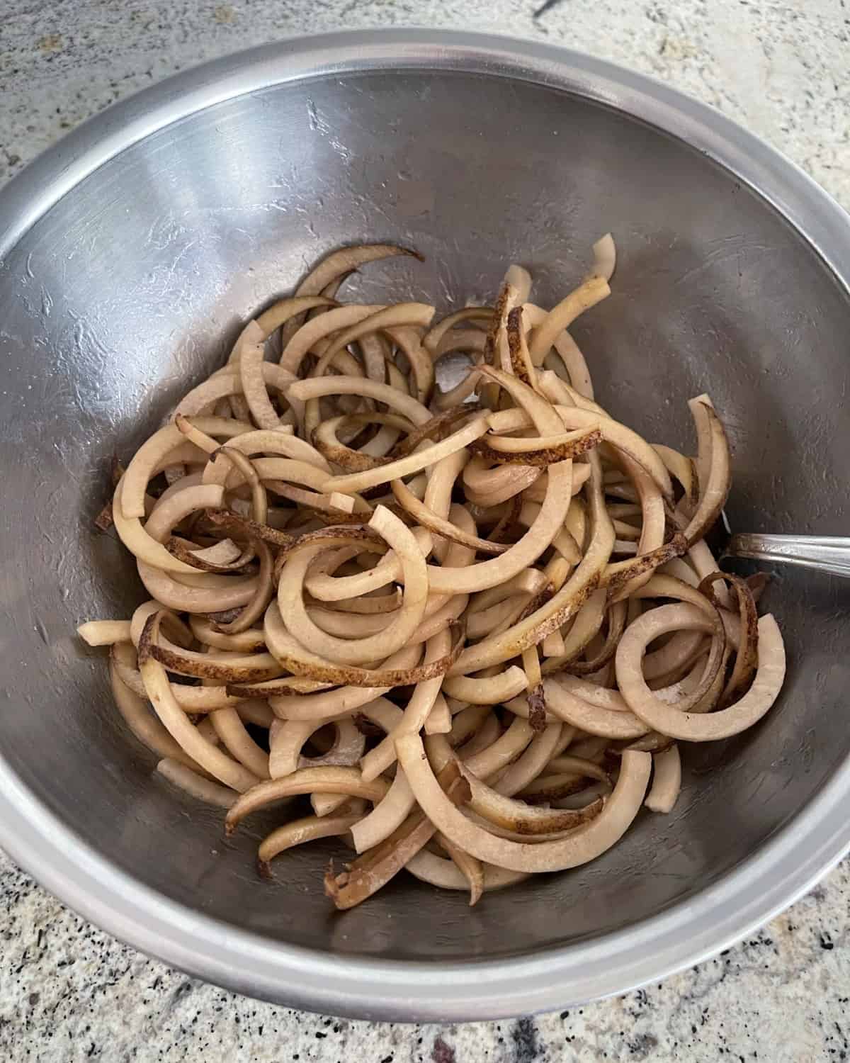 Raw uncooked curly potato spirals in mixing bowl with avocado oil and salt.