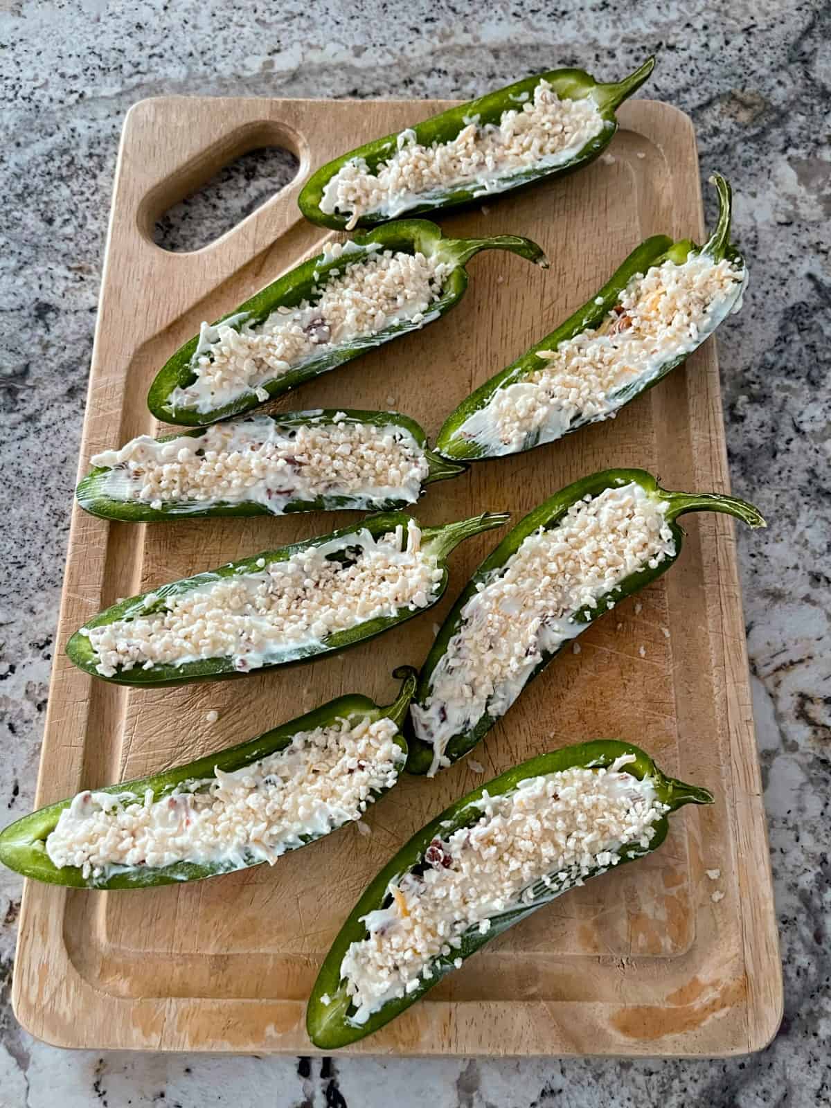 Uncooked jalapeno poppers - jalapeños cut in half and filled with a mixture of cream cheese, shredded cheese and bacon crumbles, then sprinkled with Panko breadcrumbs on wood cutting board.