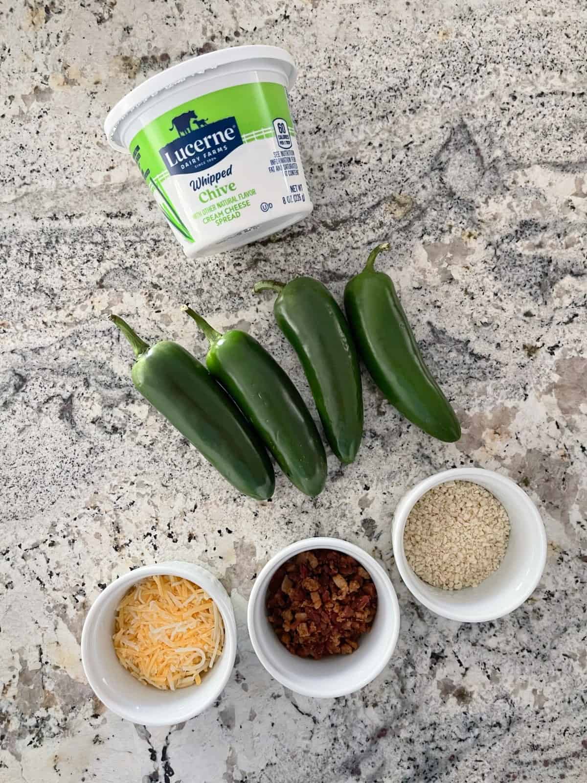 Ingredients including container of whipped cream cheese with chives, four whole jalapeno peppers, shredded cheese, bacon crumbles and panko breadcrumbs.