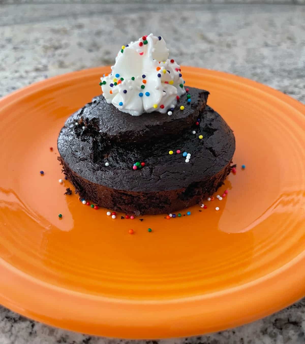 Flourless gluten-free chocolate mug cake decorated with lite whipped topping and rainbow sprinkles on small orange plate.