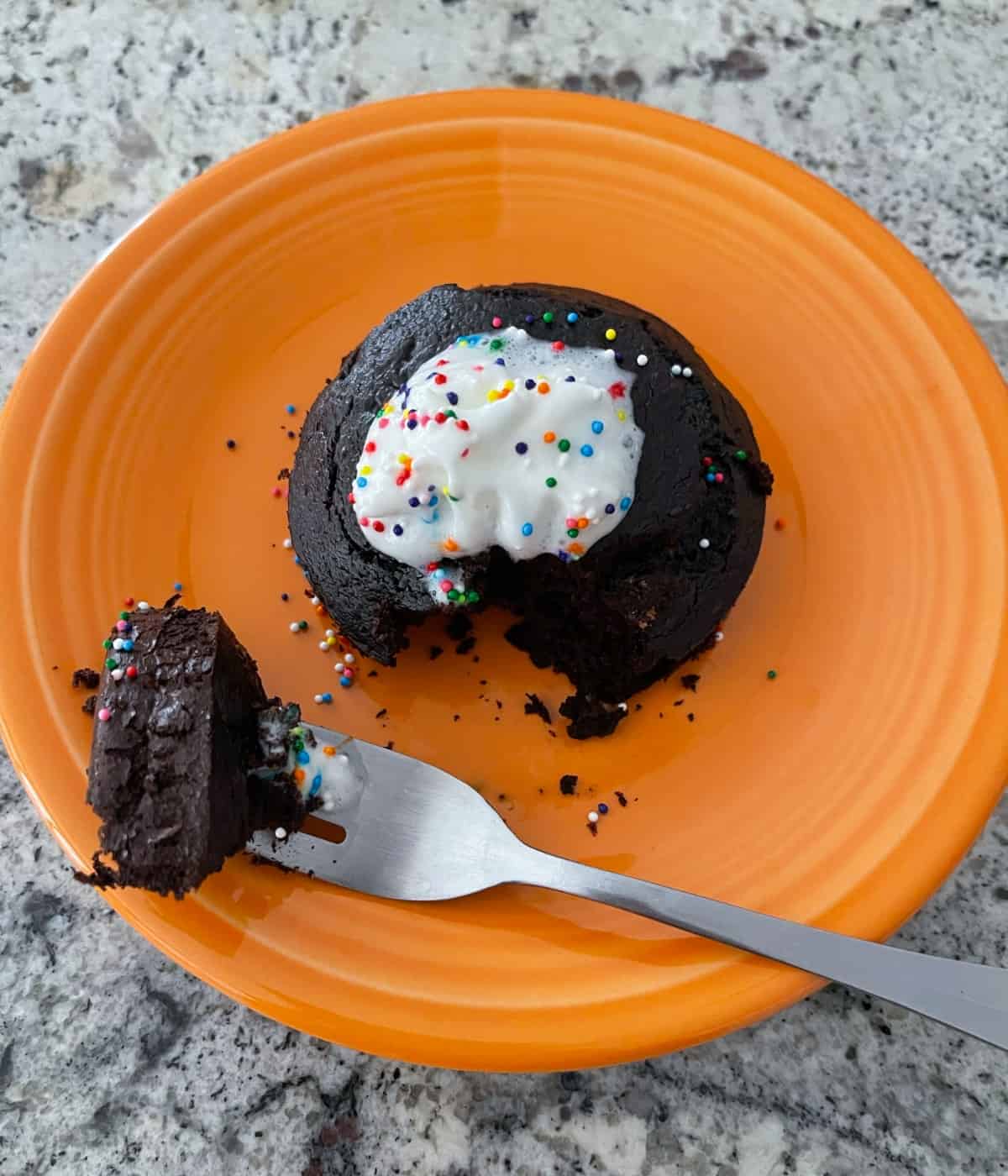 Flourless chocolate mug cake with whipped topping and rainbow sprinkles on small orange plate with fork.