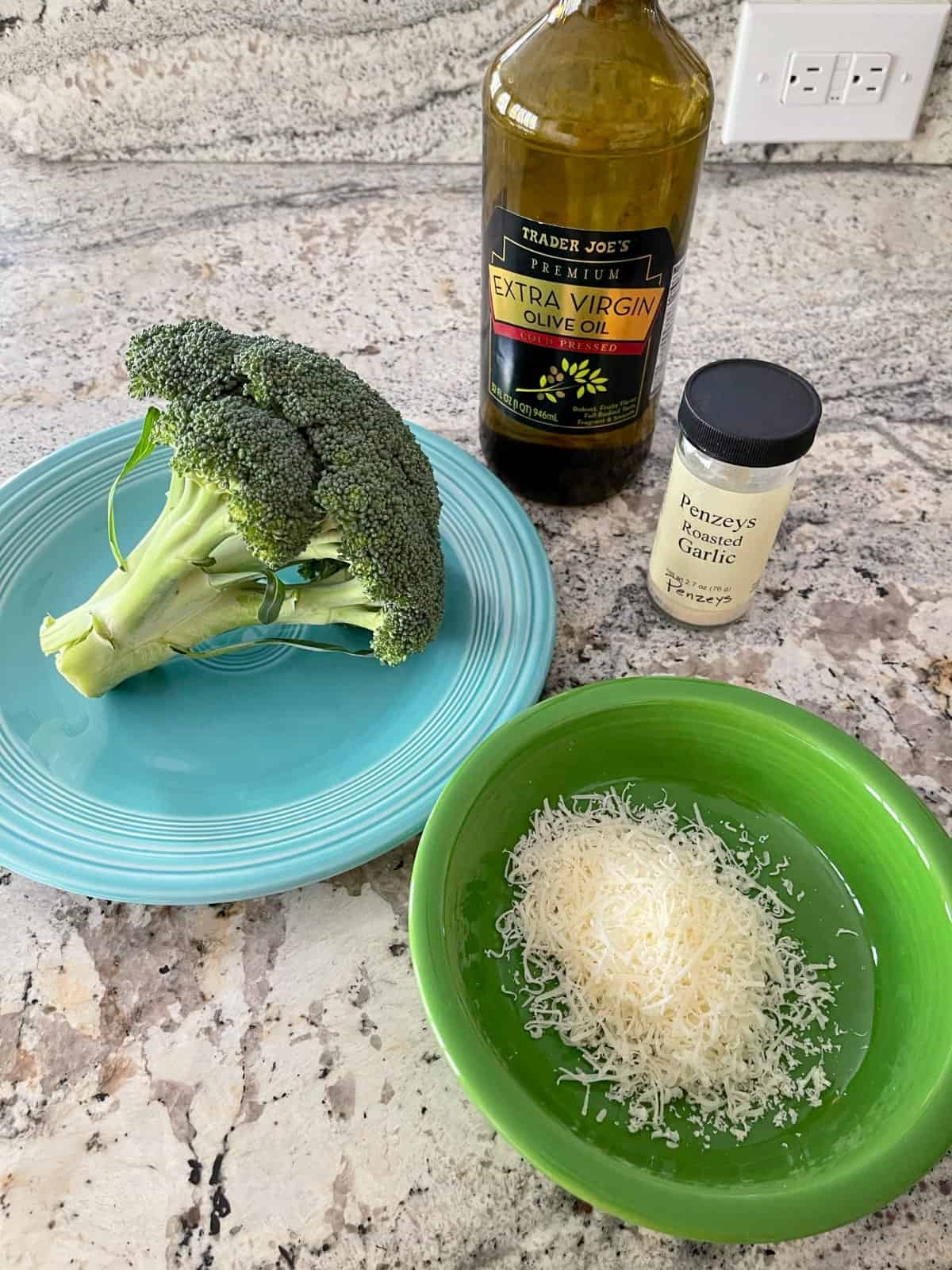 Ingredients including grated Parmesan cheese, head of broccoli, garlic powder and extra virgin olive oil on granite.