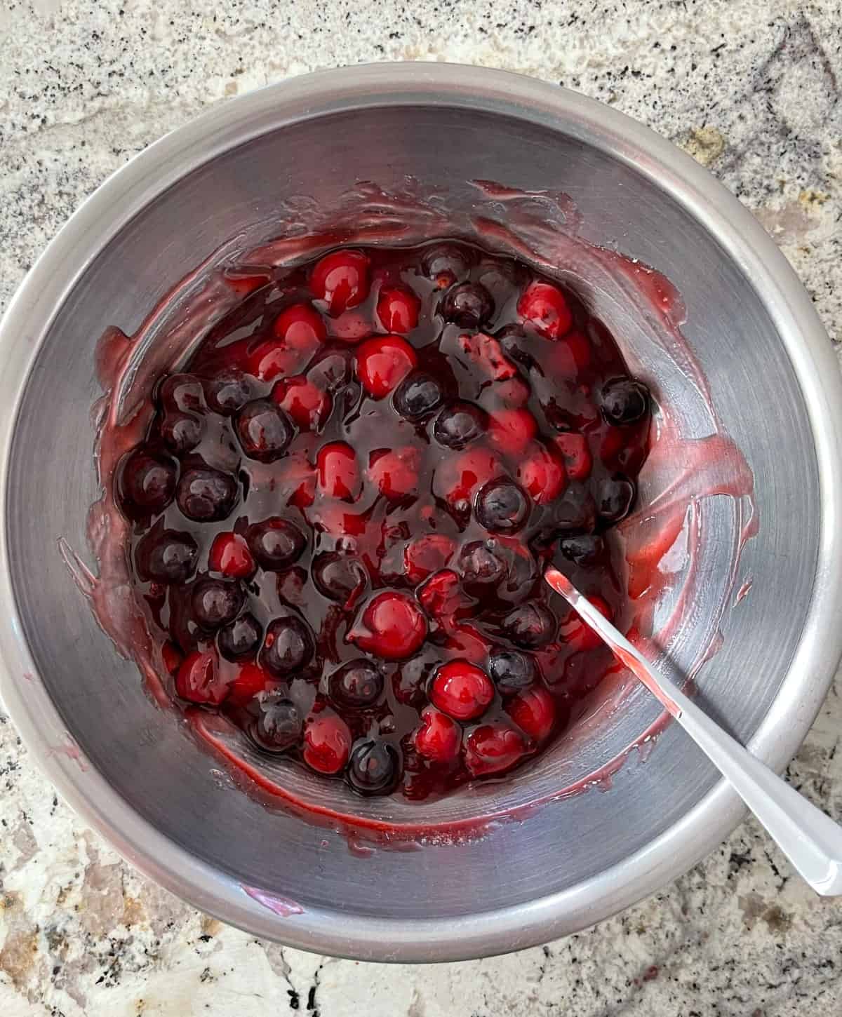 Stirring cherry pie filling, fresh blueberries, sweetener and almond extract in mixing bowl with spoon.