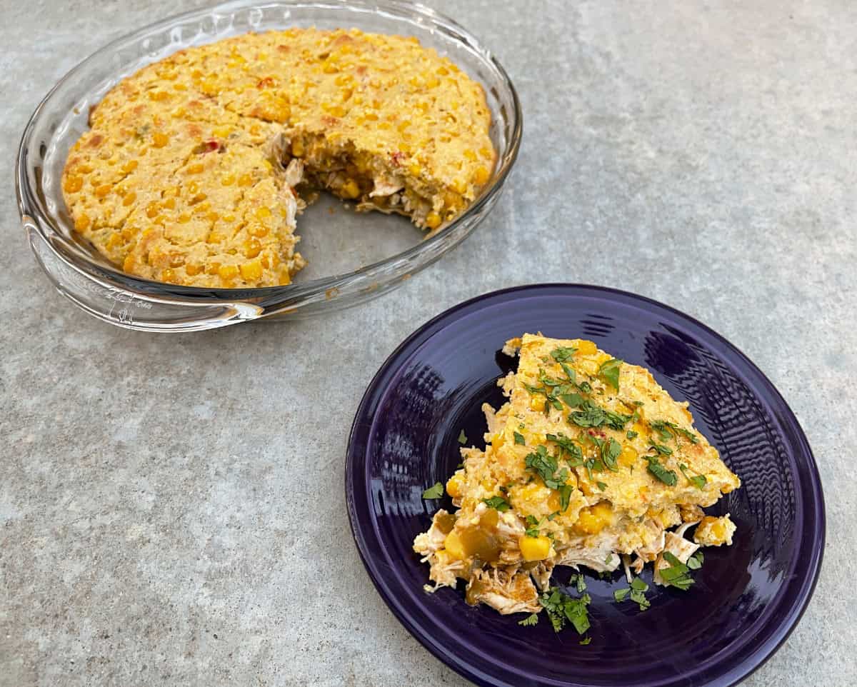 Wedge of chicken tamale pie garnished with chopped cilantro on small purple plate near chicken tamale pie in glass pie dish.