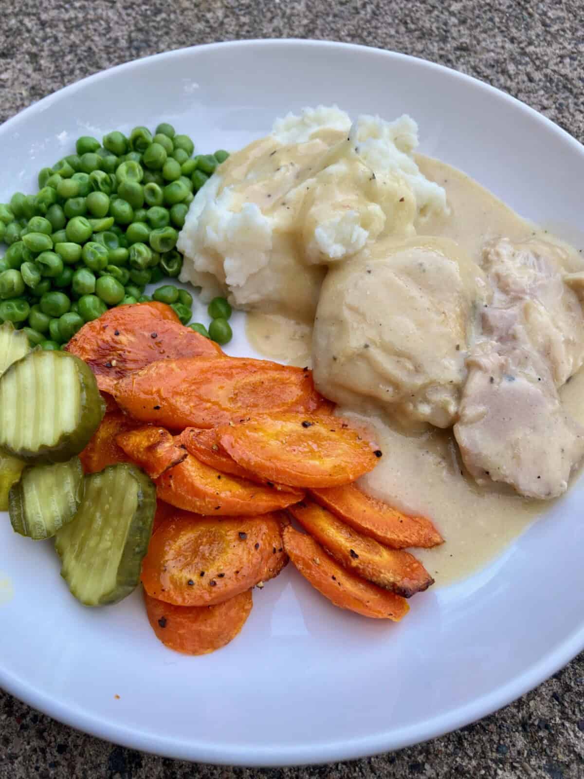 Crock pot chicken cooked in creamy soup with peas, roasted carrots and pickles on white plate