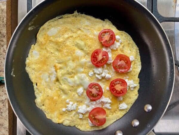 crepe style omelet in nonstick pan with feta cheese and tomatoes spread over half