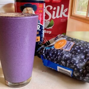 Tall full glass of blueberry Oatmeal Protein Shake surrounded by ingredients & blender