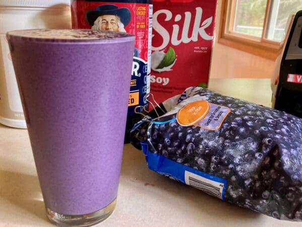 Tall full glass of blueberry Oatmeal Protein Shake surrounded by ingredients & blender