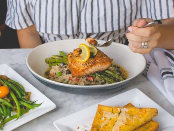 body of woman in striped blouse sitting at table with several dishes of food before her eating salmon and asparagus