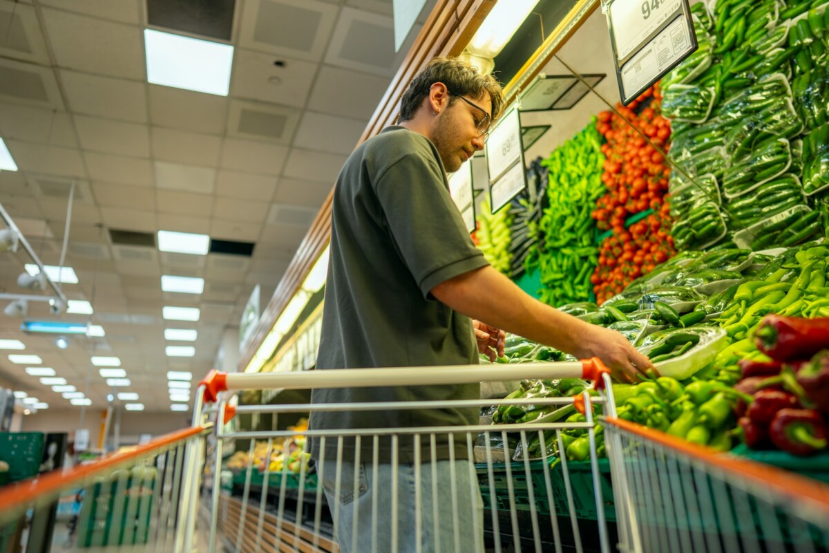 Man in vegetable aisle at grocery store with shopping cart.