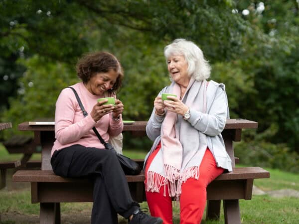 Two mature women on a park bench laughing and having coffee on