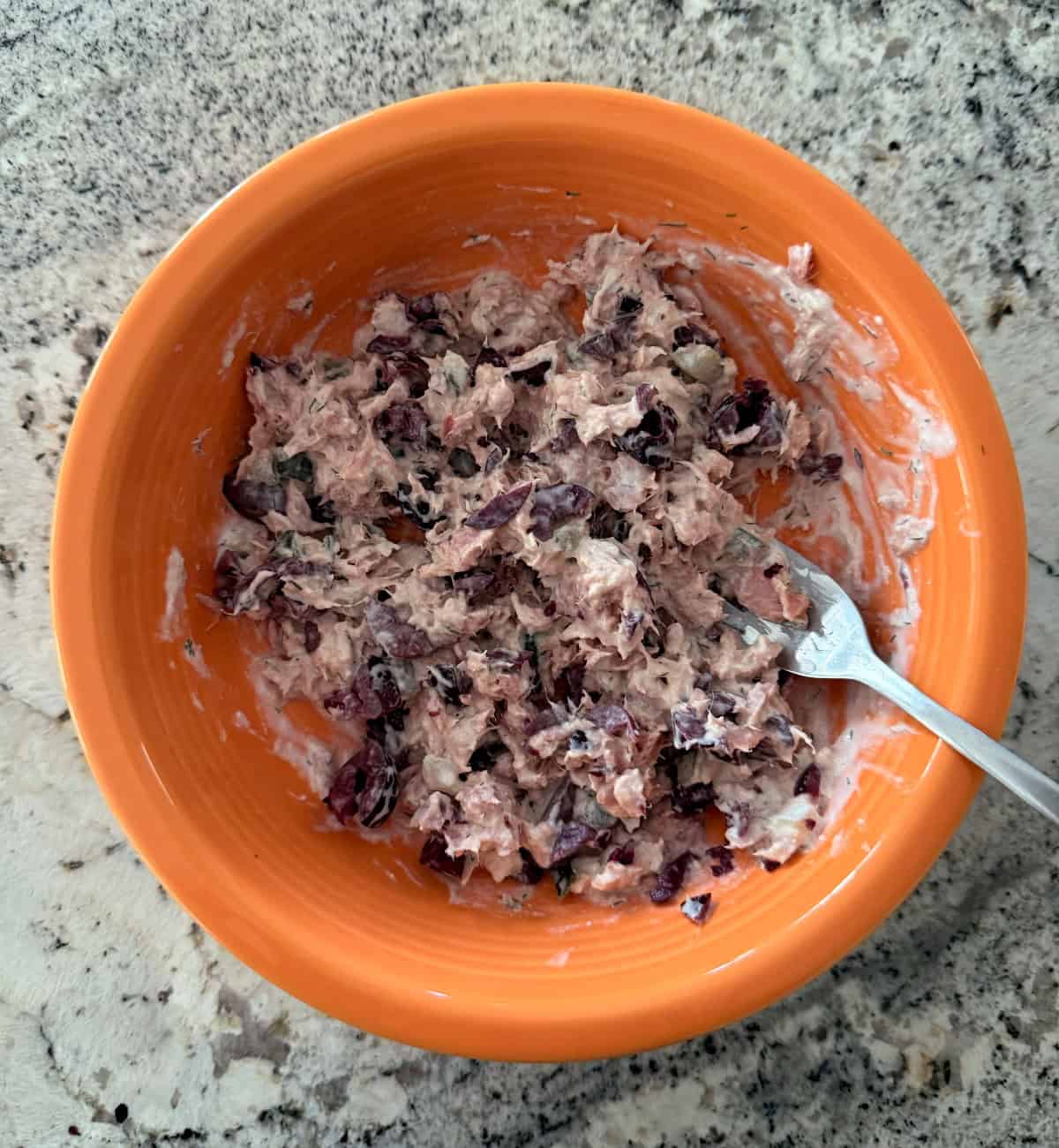 Mixing tuna with tzatziki yogurt dip and chopped Kalamata olives in orange bowl.