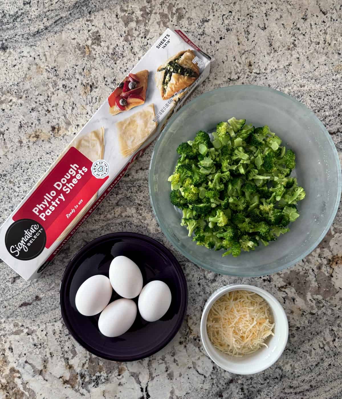 Ingredients including package of phyllo dough pastry sheets, broccoli florets, four eggs and shredded cheese.