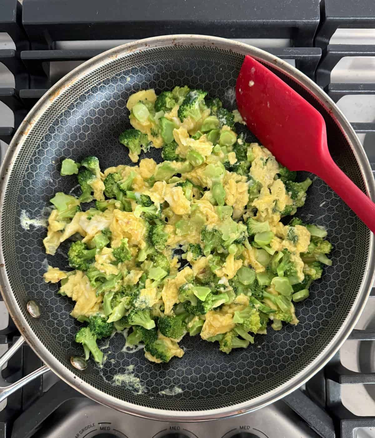 Cooking scrambled eggs with small broccoli florets in skillet with red spatula.