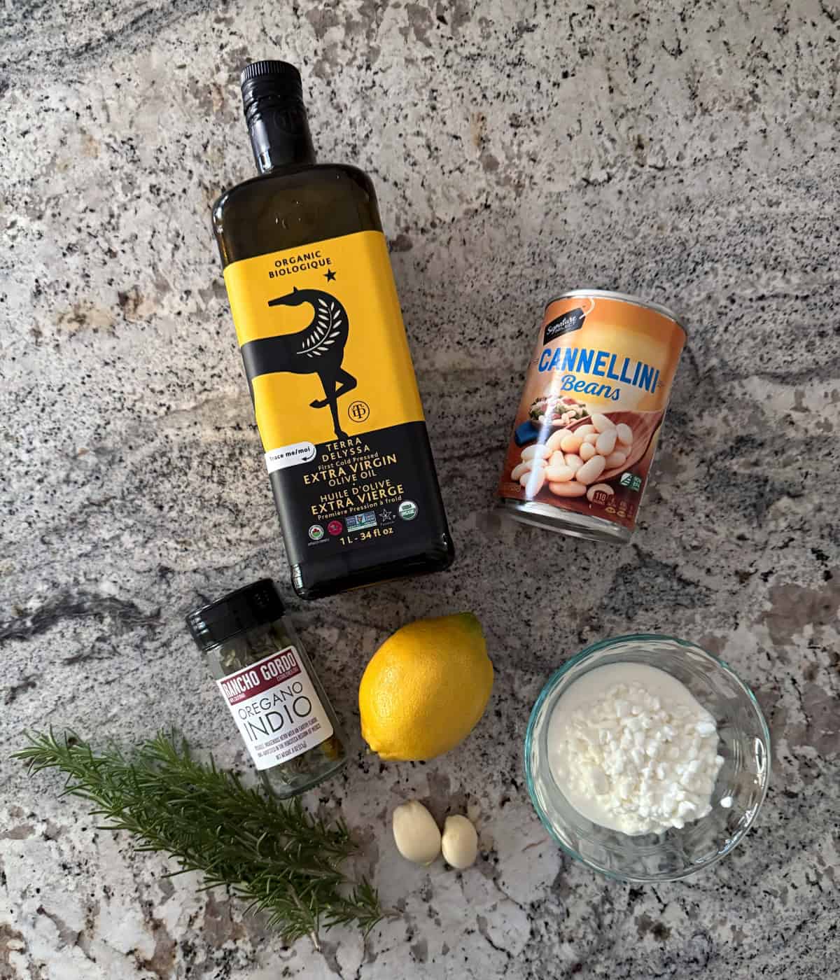 Ingredients including bottle of extra virgin olive oil, can of cannellini beans, cottage cheese, lemon, oregano, rosemary sprigs and two cloves of garlic.
