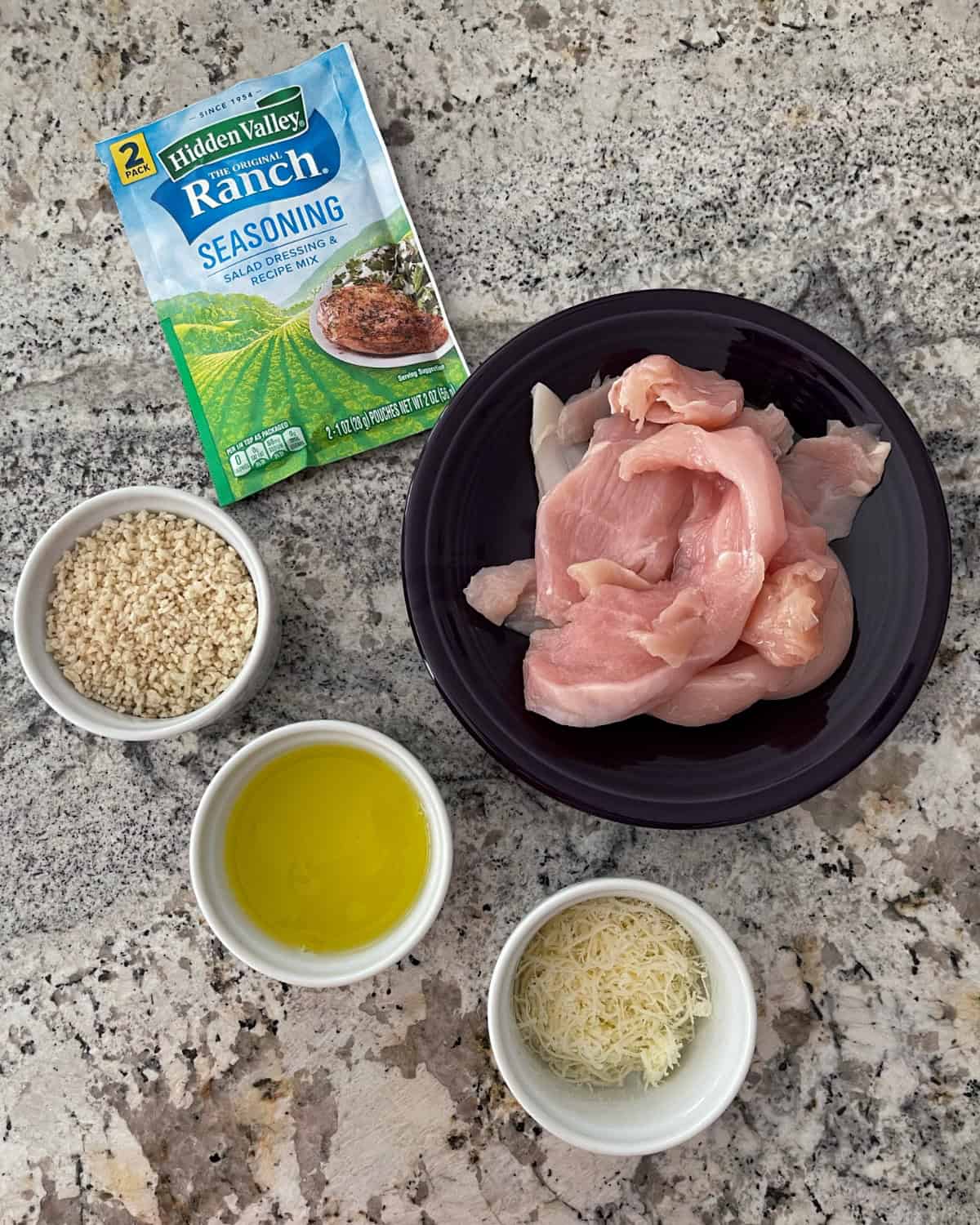 Ingredients including packet of dry ranch seasoning mix, boneless, skinless chicken breast, Panko bread crumbs, egg whites and grated Parmesan cheese on granite counter.
