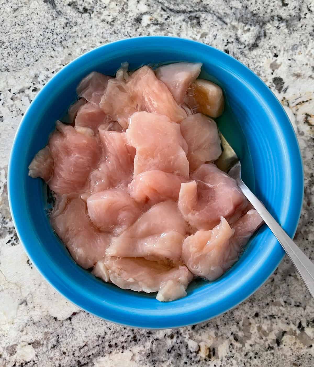 Uncooked chicken breast pieces soaking in egg whites in blue bowl.