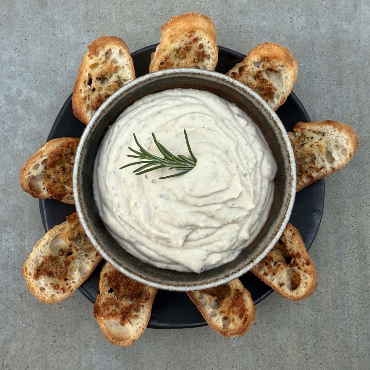Creamy Tuscan White Bean Dip in ceramic bowl with garlic crostini on brown plate.