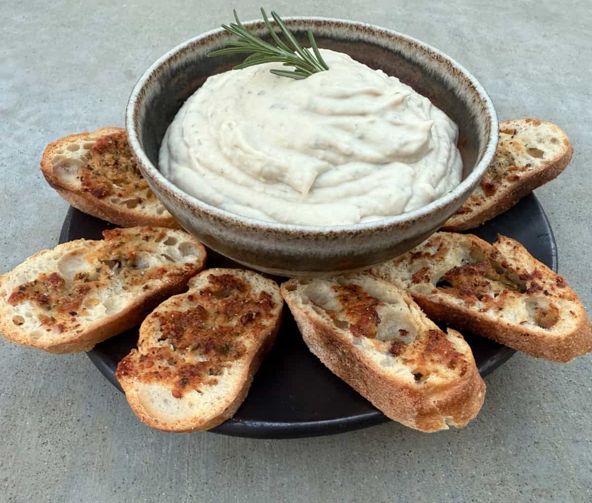 Tuscan White Bean Dip in a small bowl with a plate of garlic crostini.