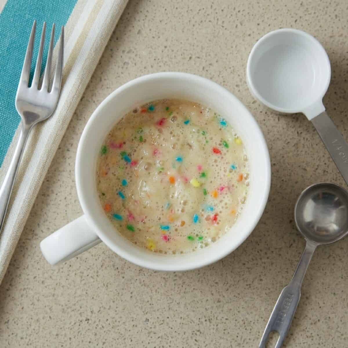 Unbaked Rainbow Mug Cake in white mug with fork and measuring spoons on kitchen counter.