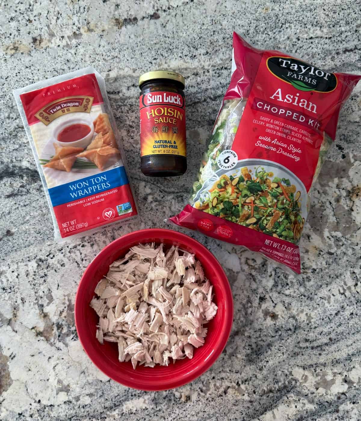 Ingredients including Asian chopped salad kit, hoisin sauce, wonton wrappers and shredded rotisserie chicken on granite counter.