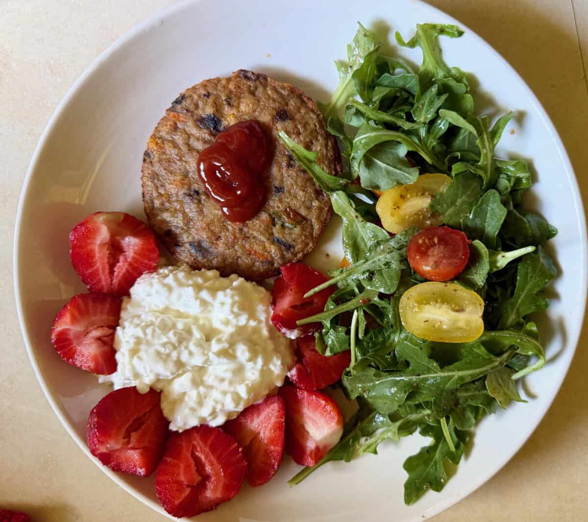 veggie burger, side salad, cottage cheese and sliced strawberries on white plate shot from above